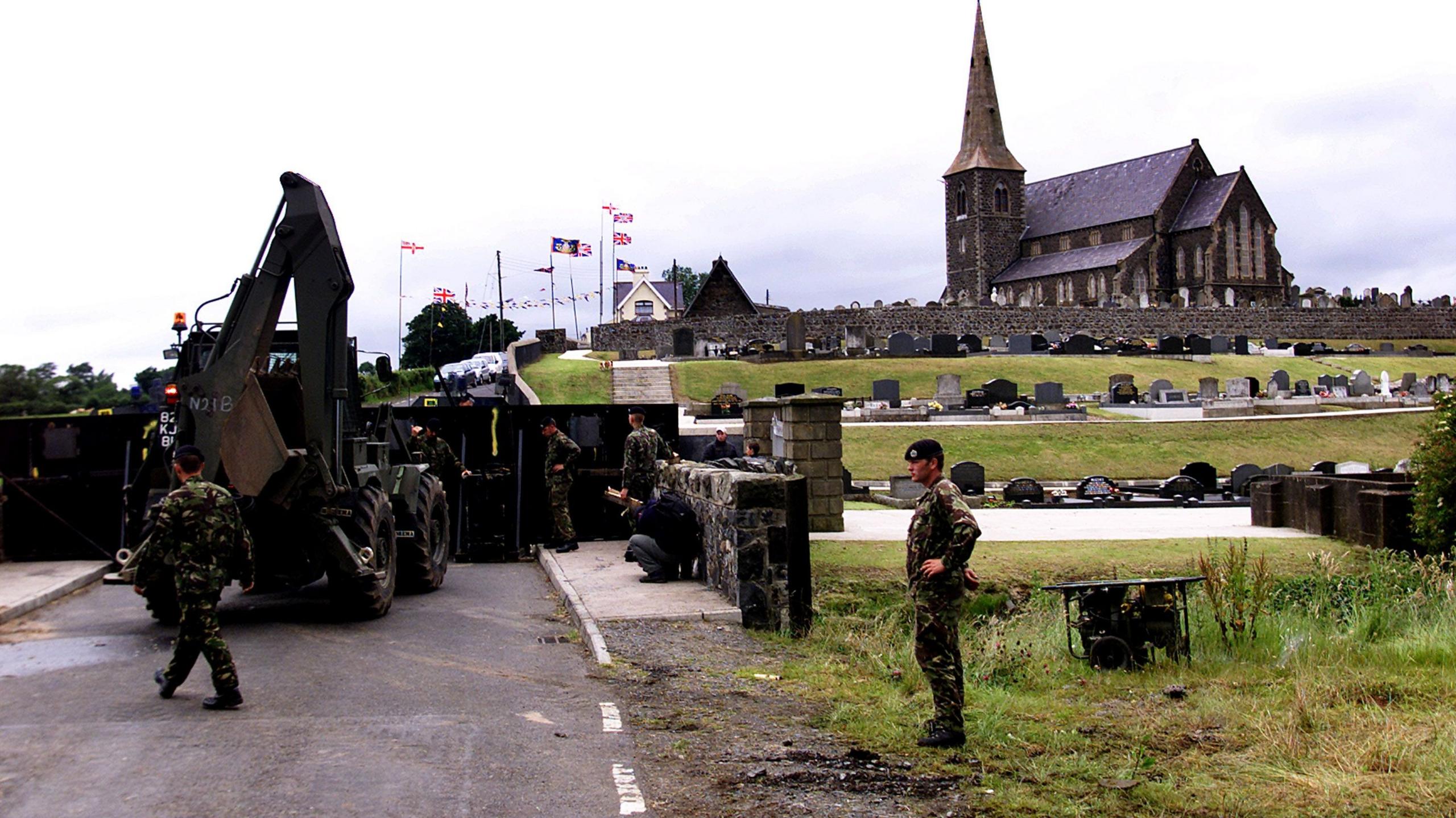 Some British soldiers are standing beside a church. A barricade is in place. Gravestones and union flags are in the background.