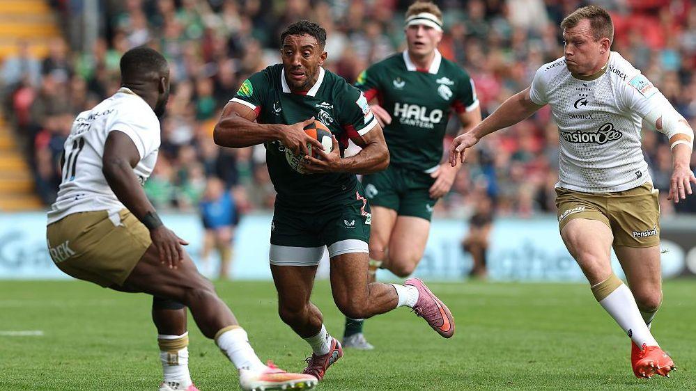 Gabriel Hamer-Webb carries the ball under his left arm as two Saracens defenders converge