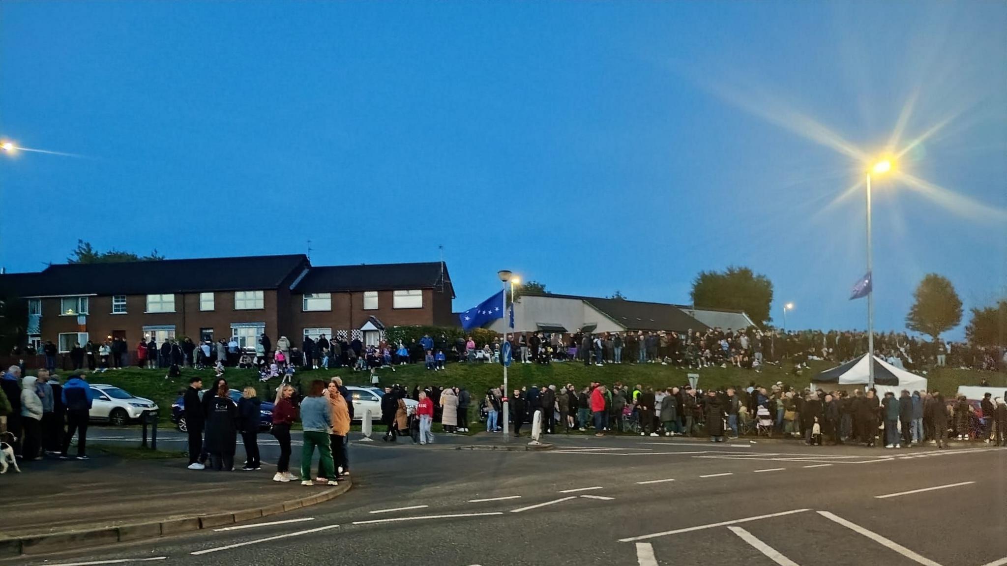 Crowds of people gather alongside a main road, with terrace houses behind them.