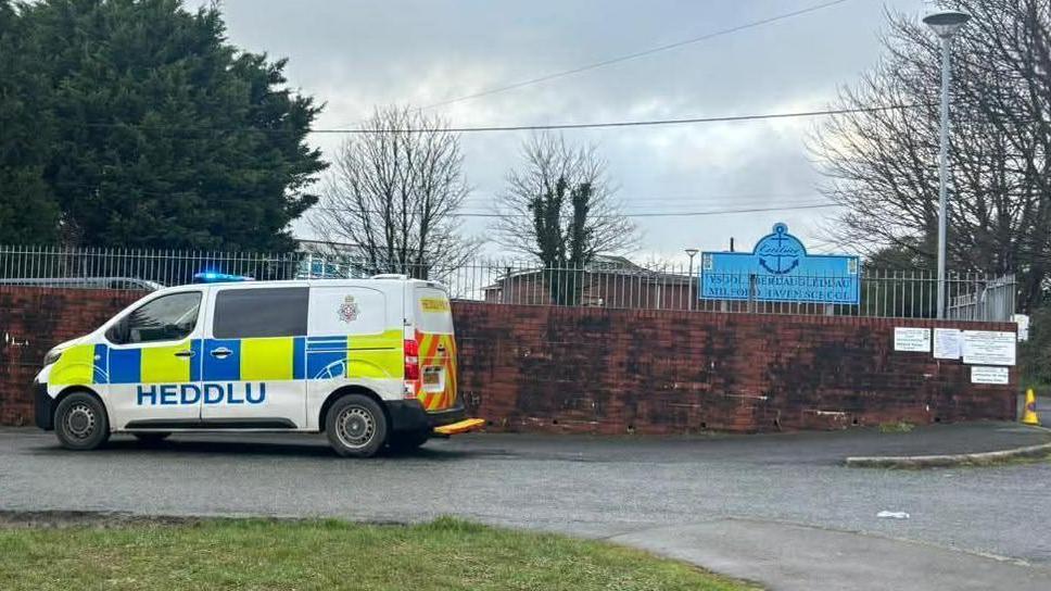 A brown school wall with a silver railing above and a police van parked outside. In the background is a blue school sign.