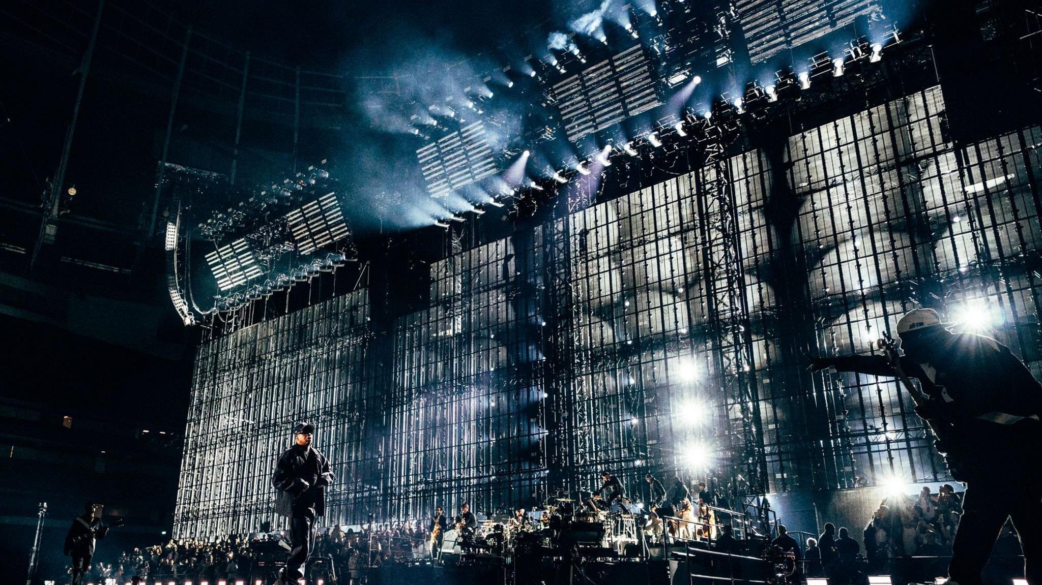 Jay-Z performs on stage in front of a large LED screen. There is an orchestra band behind him. Jay-Z wears all black.