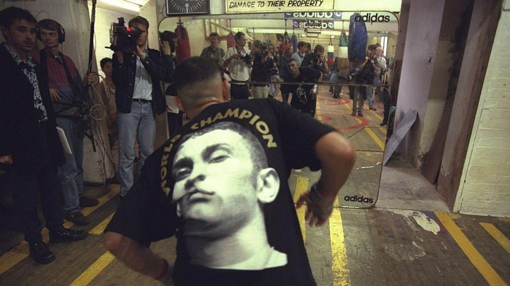 A South Asian man moving in front of a mirror while members of the media congregate to film and photograph him.
