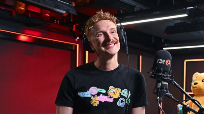 BBC Radio 2 host Owain Wyn Evans. He is wearing headphones, a black T-shirt and smiling in front of a microphone