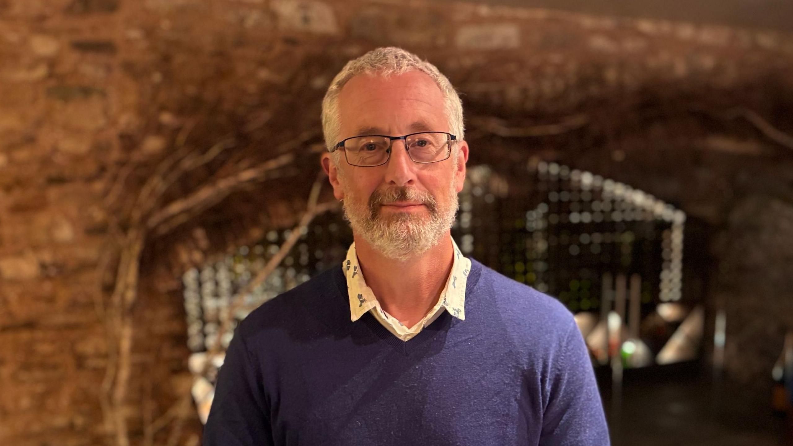 Andy is stood in a large warmly lit but shaded wine cellar. He is wearing a purple v-neck top, with an unbuttoned white shirt underneath. He wears glasses and has a short beard. Behind him is a substantial interior stone wall