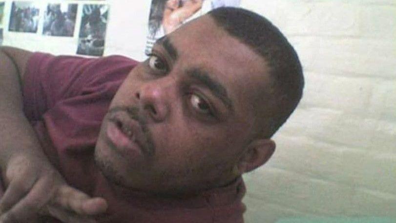 A selfie of Leroy. He is pointing at the camera, with his head and shoulders visible. He is wearing a burgundy t-shirt and has stubble and short black hair. A white brick background with posters on the wall can be seen behind him. It is a head and shoulders shot of him.