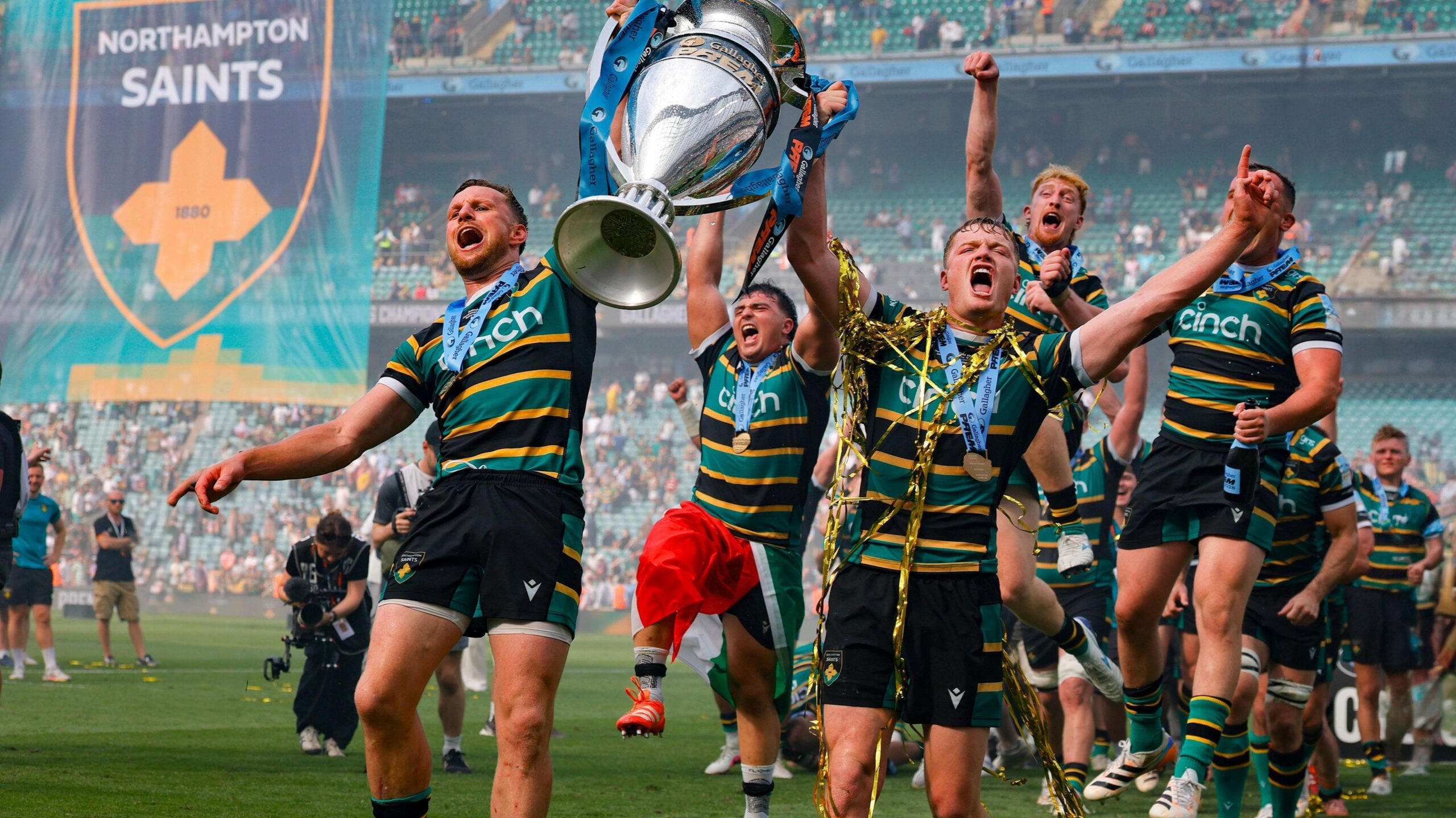 Rory Hutchinson (left) and Fin Smith hold the Prem trophy aloft between them, while on the pitch at Allianz Stadium after the full-time whistle of the Prem final. Northampton players run and jump behind them