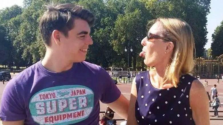 Oliver McGowan, wearing a purple t-shirt with a logo, is standing outside Buckingham Palace. He is starring at his mother Paula McGowan who is standing next to him and wearing a dark blue polka dot dress and sunglasses. She is playfully sticking her tongue out at her son and he is laughing.