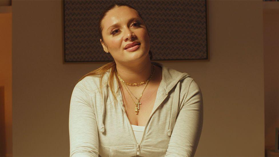 Medium close-up of Violet seated indoors under warm lighting, wearing a light-coloured zip-up hoodie over a white top. Several necklaces, including a cross pendant, are visible. She is facing slightly away from the camera, with a framed patterned artwork on the wall behind her and a softly lit interior background.