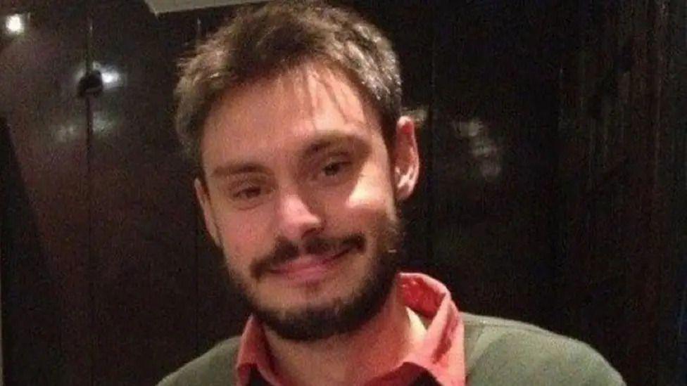 Giulio Regeni smiling at the camera wearing a pink collared shirt with a green jumper