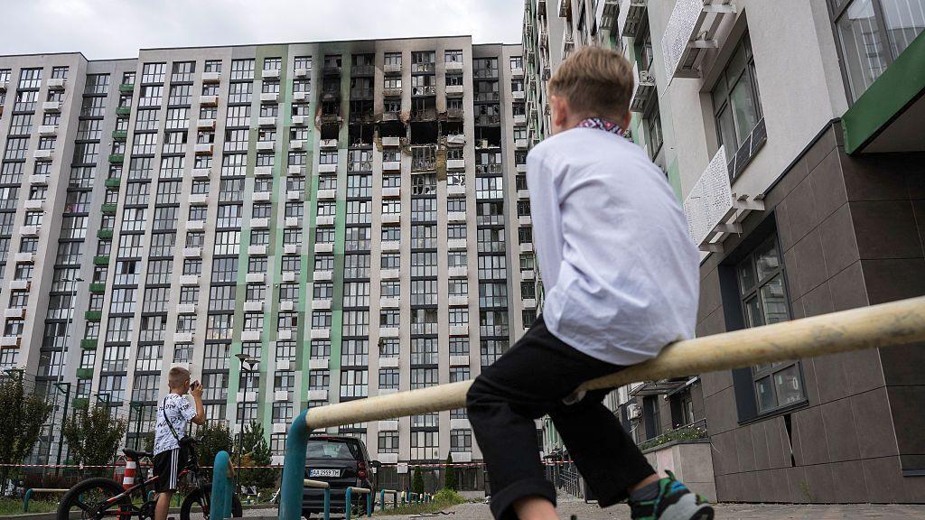 A little boy in a white embroidered shirt and black trousers looks at a building damaged in a Russian air attack, in Kyiv