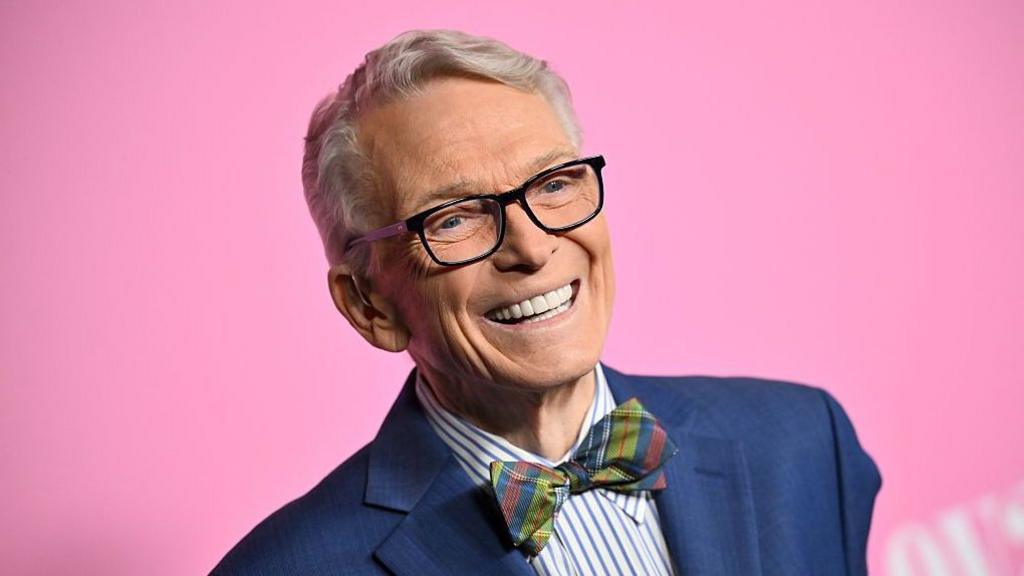 Bob Mackie wears a navy blue blazer, blue and white striped shirt and a multi-coloured tartain style bow-tie. He's smiling and there's a bright pink background.