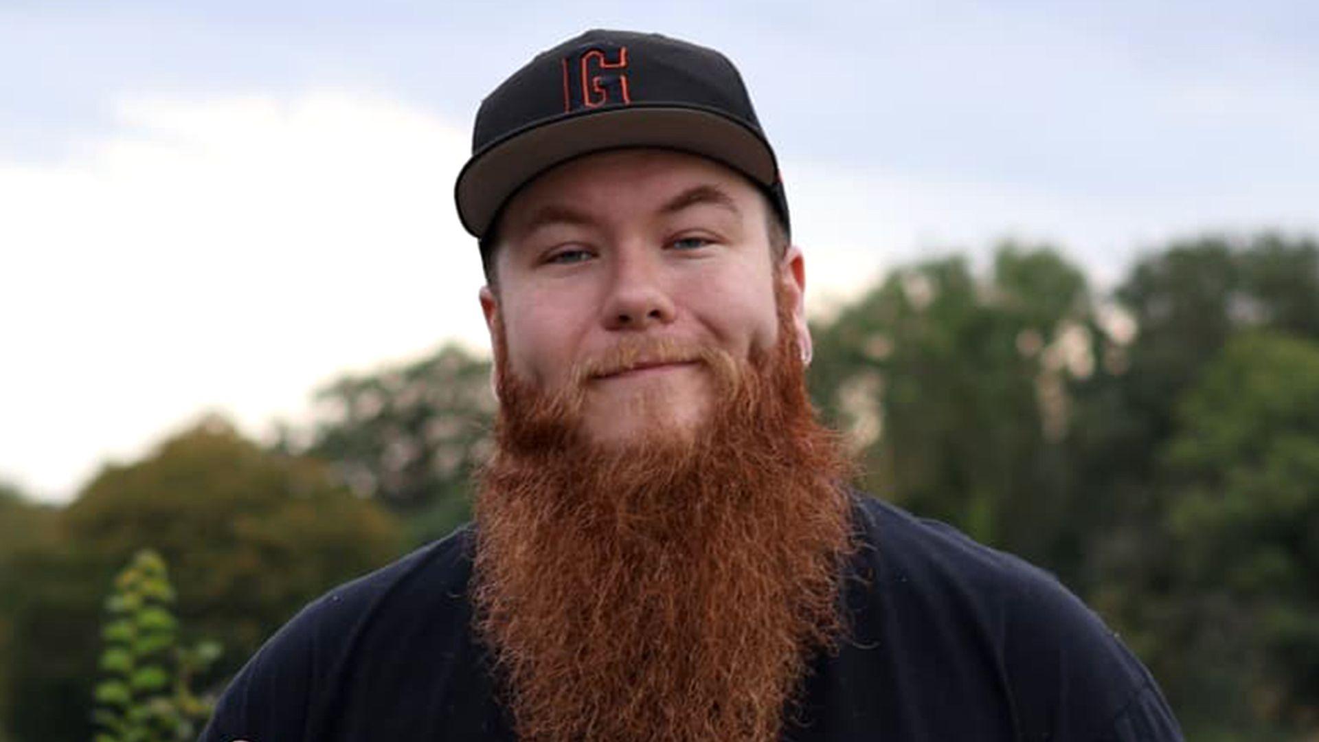 Luke has a long reddish‑brown beard and stands outdoors wearing a black baseball cap with an orange “G” and a dark shirt, set against green trees and a cloudy sky.