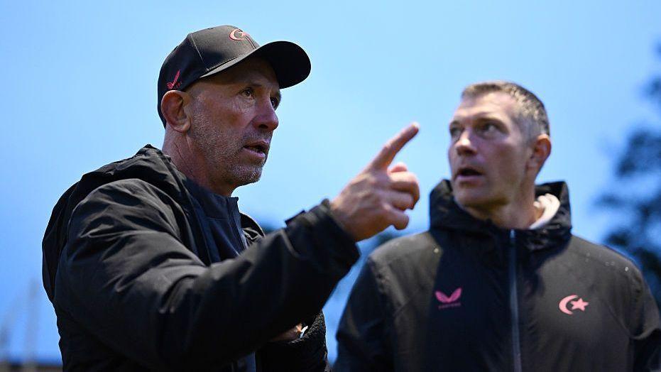 Brendan Venter (left) points his finger as he talks. He is wearing a black cap, a black coat.