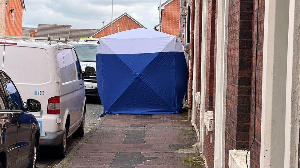 Man arrested on suspicion of murder after two people found dead at house Blue and white forensic tent on a residential pavement between parked vans and a row of terraced houses.