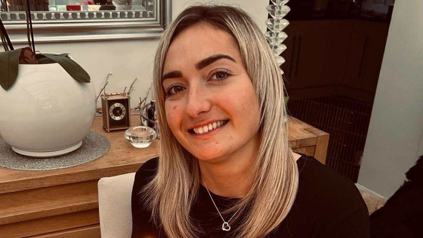 Maddy Cusack pictured smiling at home. She has long blonde hair worn loose and wears a silver heart necklace over a black top. She's pictured inside, in front of a side board with ornaments and plants