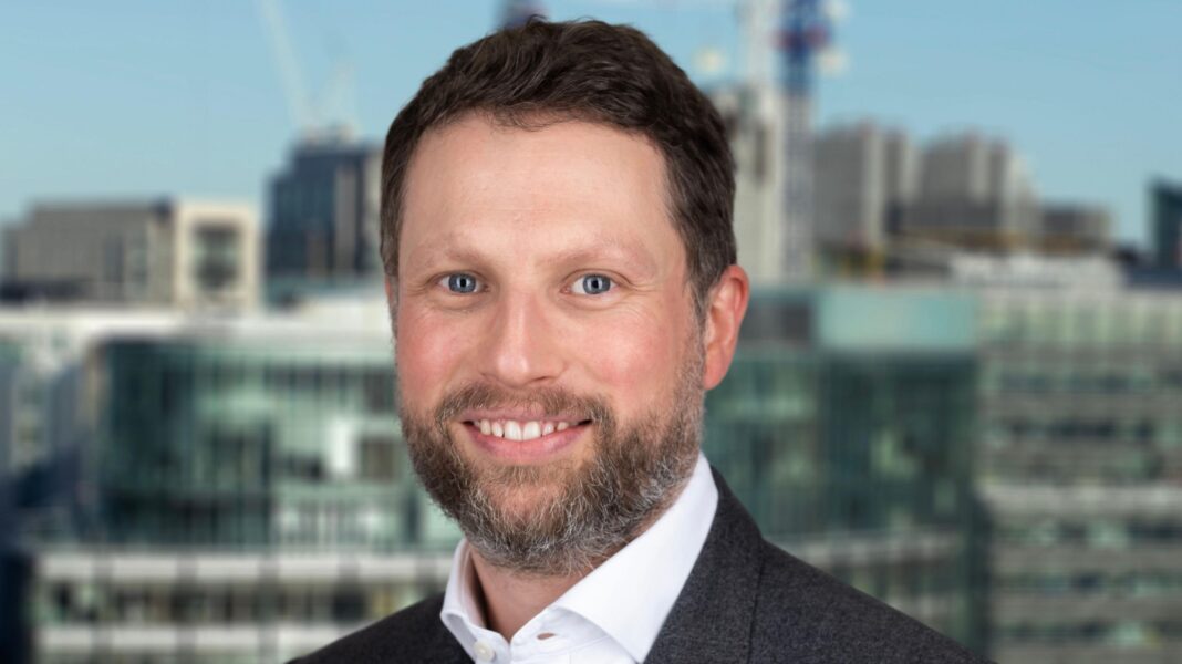 A headshot of a smiling man looking straight at the camera. He is wearing a grey suit jacket and a white shirt with an unbuttoned collar. Behind him it's possible to see a blurred image of the city of London skyline