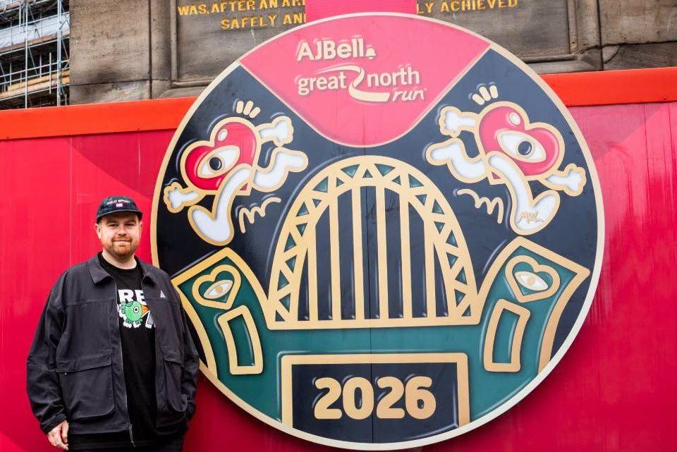 Alex Mulholland is standing beside a large mock-up of the medal and smiling at the camera. The black, green and red cartoon design features the zig-zagged metal features of the Tyne Bridge in the middle, with two heart-shaped characters running on either side.