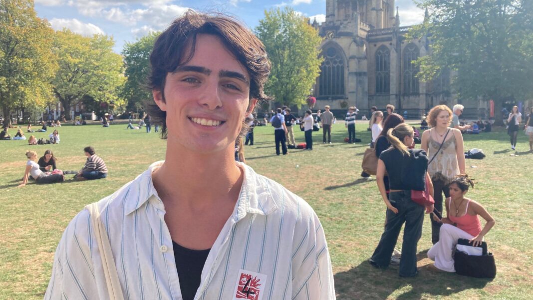 Joey Lush wearing a black T-shirt underneath a white collared shirt with blue stripes and a white tote bag on his shoulder. He has brown hair and thick eyebrows and he is smiling at the camera. It is a bright sunny day and there are lots of people standing and sitting on the grass on College Green, with the cathedral behind them.