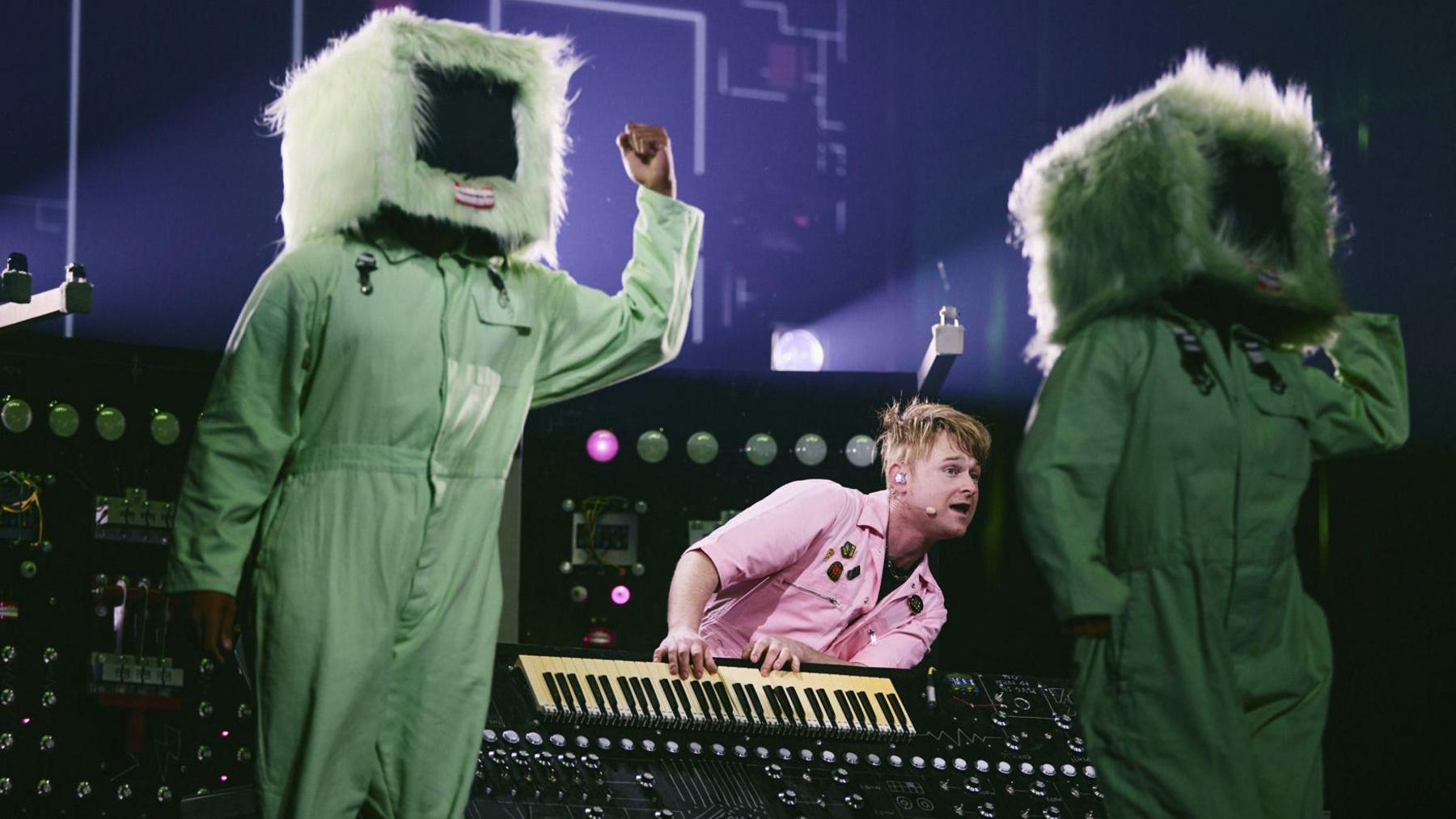 A man in a pink shirt playing a keyboard. On either side of him are two people, dressed in a green jumpsuit and a television prop over their heads.