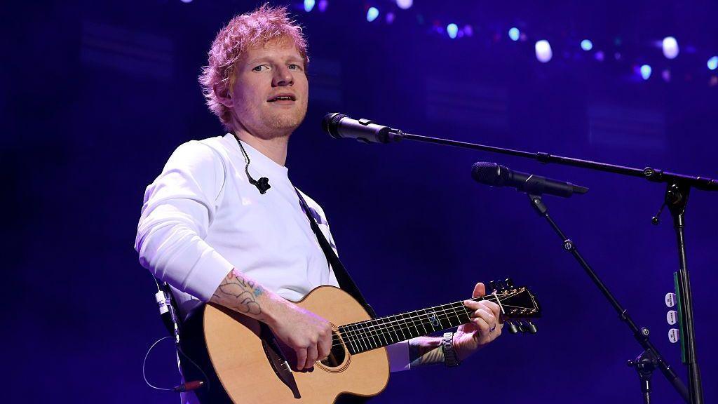 Ed Sheeran, in a long sleeved white T-shirt, holds a guitar and sings into a microphone. He is looking slightly off to one side.