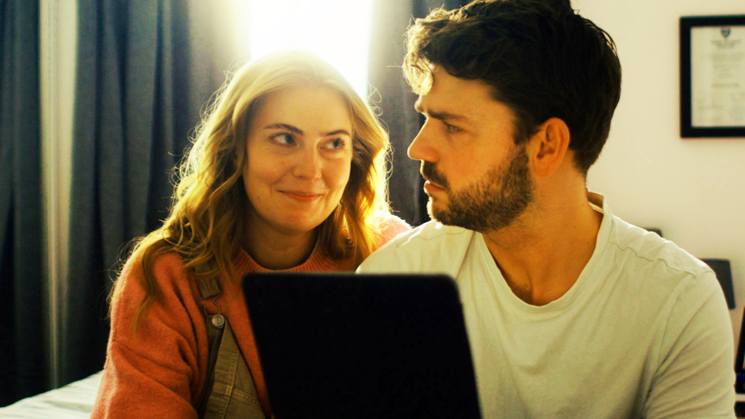 A man and woman sit closely together in a softly lit room, looking at each other over the top of an open laptop. Bright daylight streams through a window behind them, framed by dark curtains. The woman is wearing a pink jumper and dungarees, while the man is wearing a white T-shirt and has dark hair and a beard.