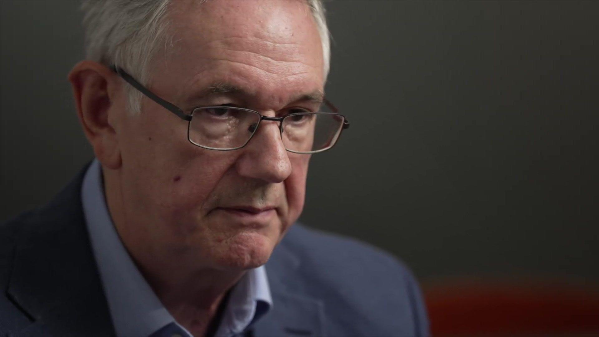 A man, wearing black, square-frame, glasses looks down. His face is stern. He has grey hair and a blue suit jacket and blue shirt on. The background is blurred.