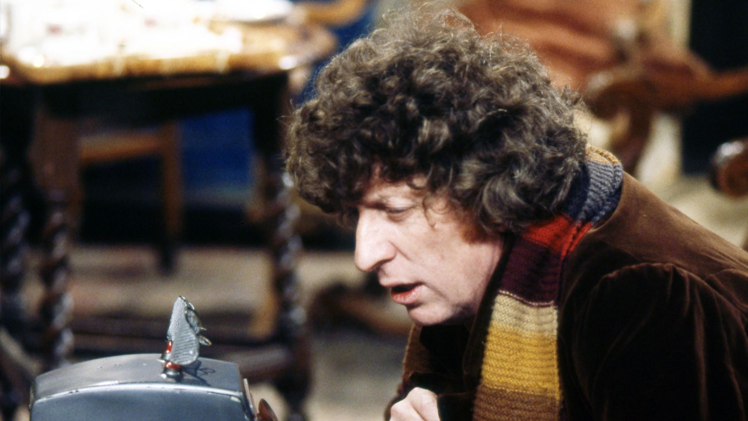 Tom Baker has curly brown hair. He is wearing a brown coat and a multi-coloured long scarf. He is sitting on the right and looking towards a silver device.