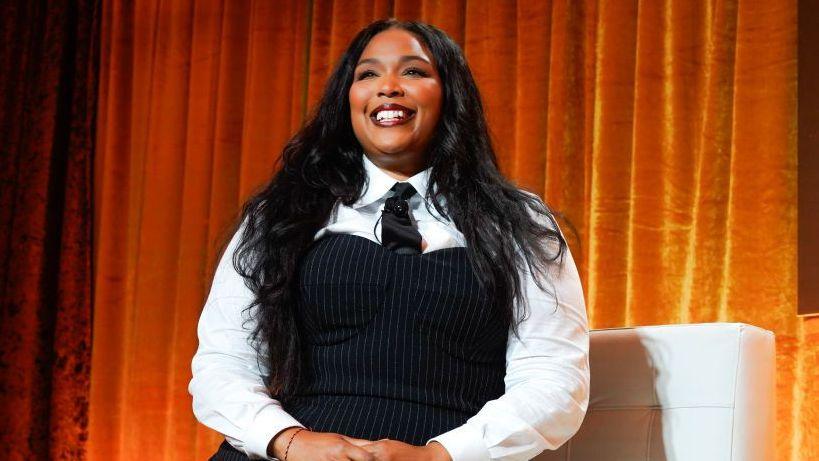 Lizzo sits on a white sofa against an orange curtain and faces out to a crowd with a smile. She wears a white shirt with a pinstrip corset style dress and black tie. Her black hair sits on her shoulders.