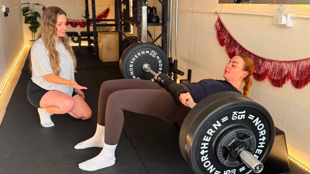Treadmills to deadlifts: Why Gen Z are swapping cardio for strength training Antonia, on the left, wearing a grey t-shirt and black shorts, is talking to a young female client while she is doing hip thrusts using heavy weights on a bar which is lying across her hips