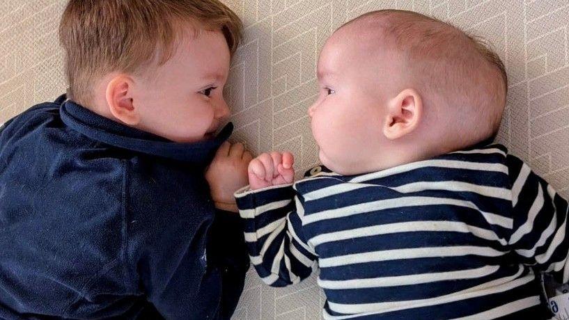 A little boy is laid on the floor on his front. He is looking at his baby brother who is laid on his  back next to him.