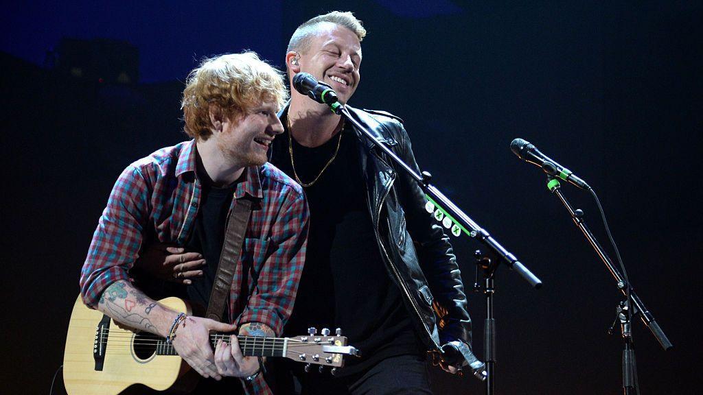 Sheeran holding guitar and smiling over at Macklemore who is also smiling