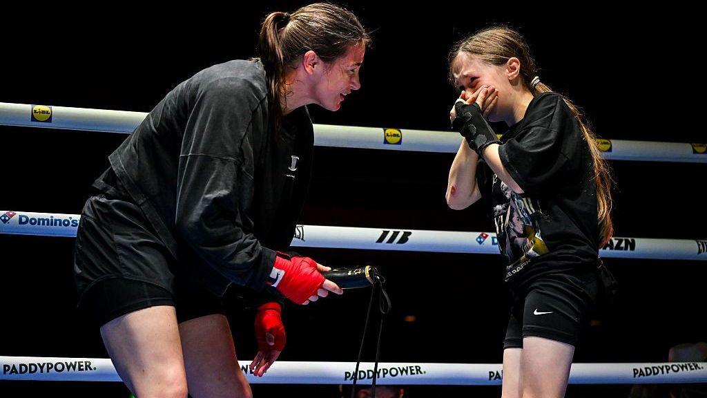 Katie Taylor crouches down and holds out a glove as Clodagh Keating holds her hands over her mouth in shock
