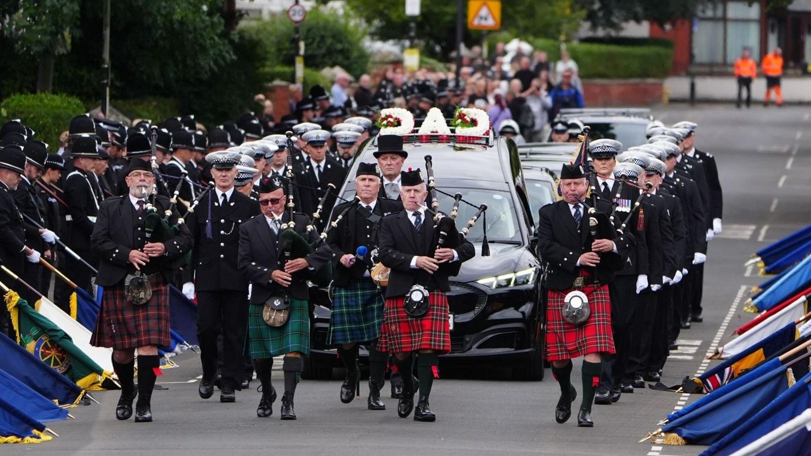 The funeral procession which is being led by five men in different coloured kilts playing bagpipes. Behind them is a black hearse with a white, red and green floral tribute on top of it which spells out 'Dad'. Uniformed police officers in black suits and white hats walk alongside the vehicle. Police officers in black uniform and black hats line the street on either side with different coloured flags resting on the road in front of them. In the distance is a crowd of people on the left who are out of focus.