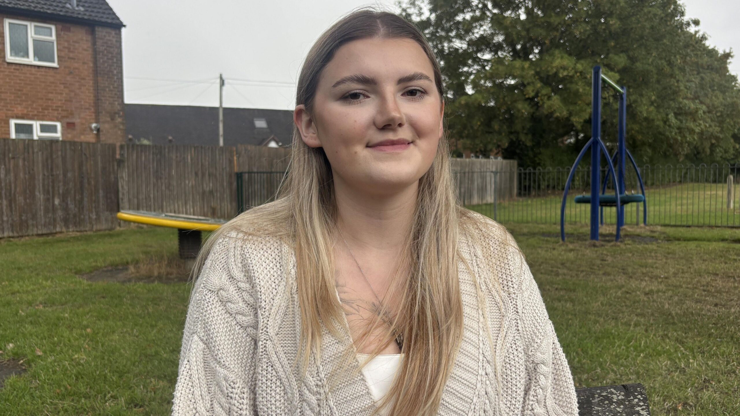 Caeila Dorsett, Morgan's sister, wearing a cream knitted cardigan over a white top. She has long blonde hair which is slightly darker at the roots, and is sitting on a park bench, smiling at the camera.