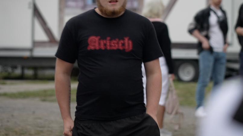The torso of a man wearing a black t-shirt which says 'Arisch', German for Aryan written in a red Gothic-style font.