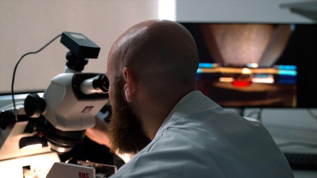 How the brain of a fly may help explain human behaviour A close, cinematic view from behind of a bald, bearded scientist in a pale blue laboratory coat. He leans forward to look through a large white microscope, its black eyepiece pressed near his face. A small camera-like device is mounted on top, with a cable trailing away. Bright light illuminates the microscope’s sample stage and the man’s shoulders, while the surrounding laboratory is dim. In the background, a large monitor displays a blurred, magnified image in warm brown tones, crossed by sharp horizontal streaks of blue, white, orange and red light