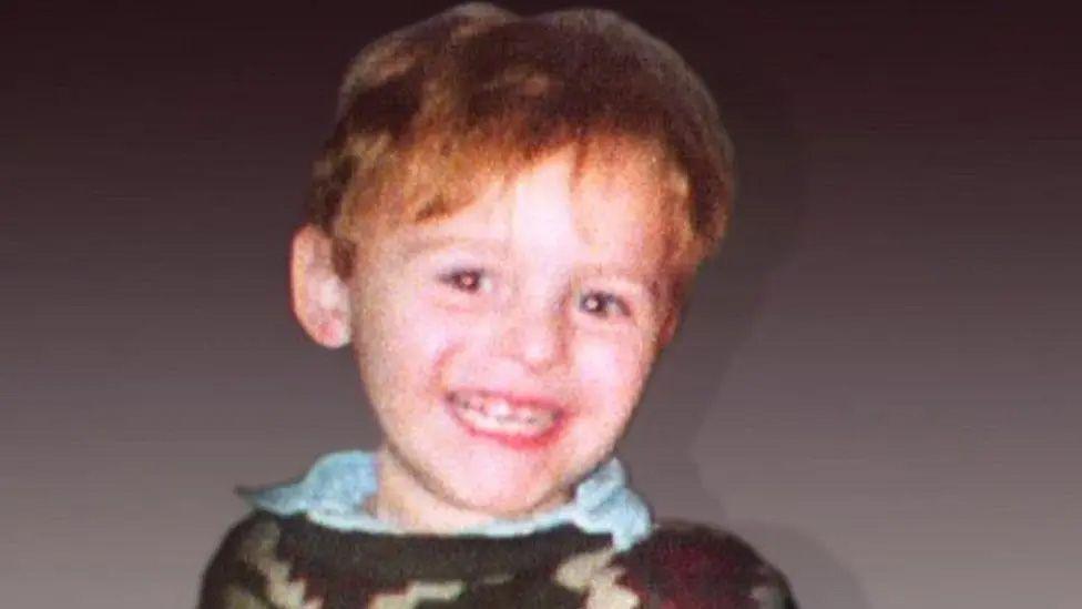 James Bulger is wearing a brown jumper over a blue shirt and is smiling at the camera.