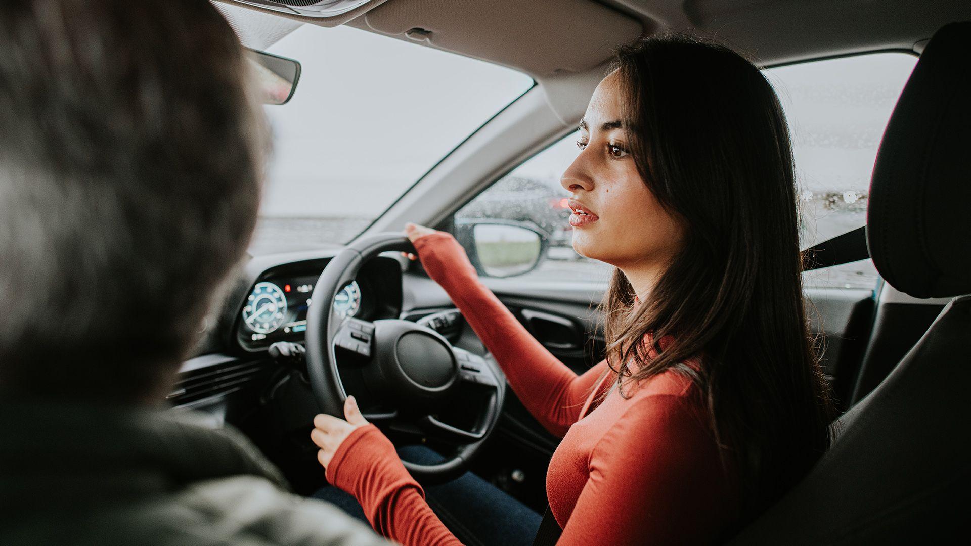 Stock image shows a young person turning a steering wheel as they reverse in a car while an instructor (out of frame) sits in the passenger seat.
