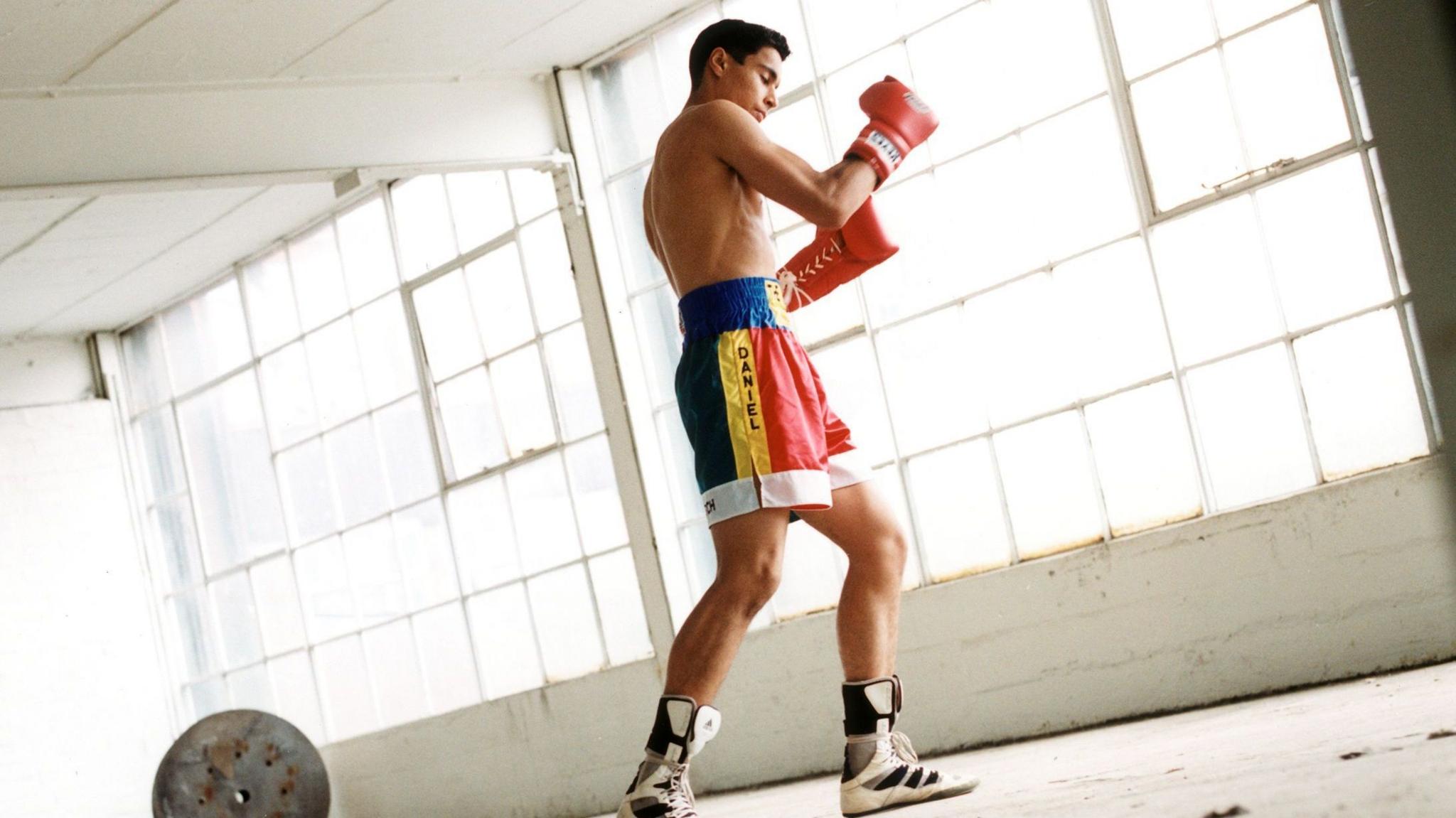 A young boy shadow boxed in what looks like an empty warehouse with bright natural light shining into the room. He is wearing blue, yellow and red shorts and red gloves.