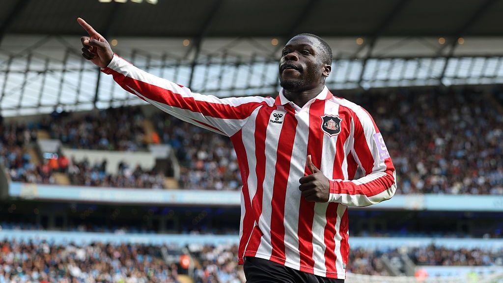 Brian Brobbey, wearing Sunderland's red-and-white-striped kit, celebrates his goal against Manchester City by pointing to the crowd