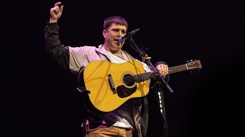 Rowe playing a guitar on stage in front of microphone, with one arm raised