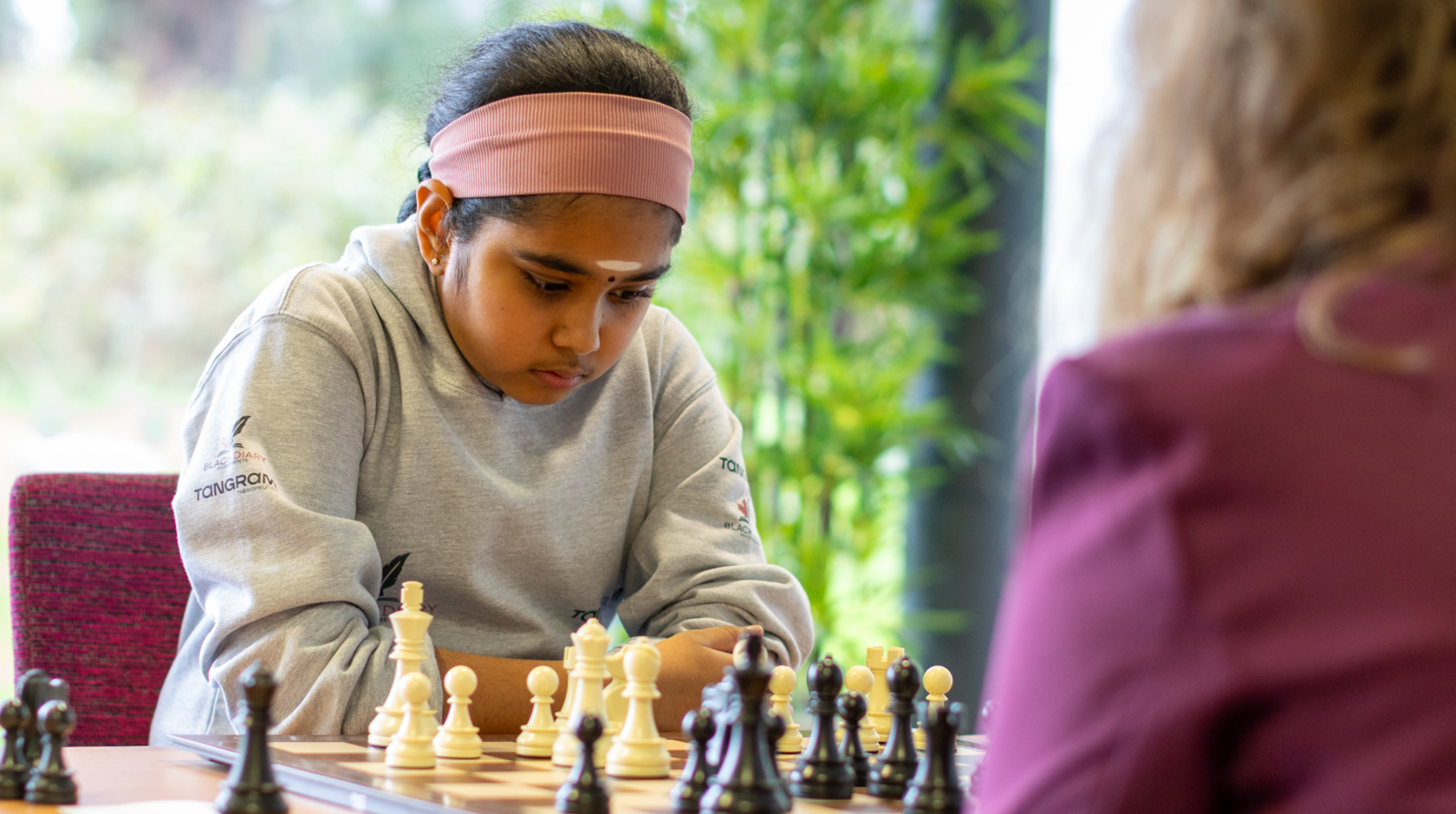 Bodhana wearing a grey sweater, and with a pink headband, stares with deep concentration at a chess set. She is playing against an adult woman.