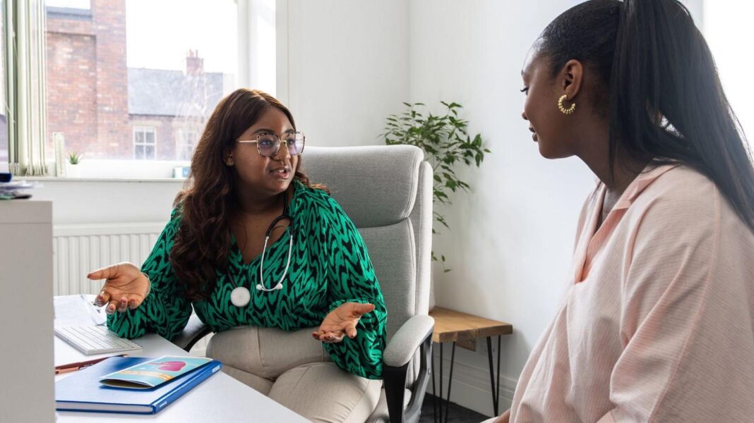 A woman in a doctor's office. The doctor is wearing a stethoscope and talking to the patient.