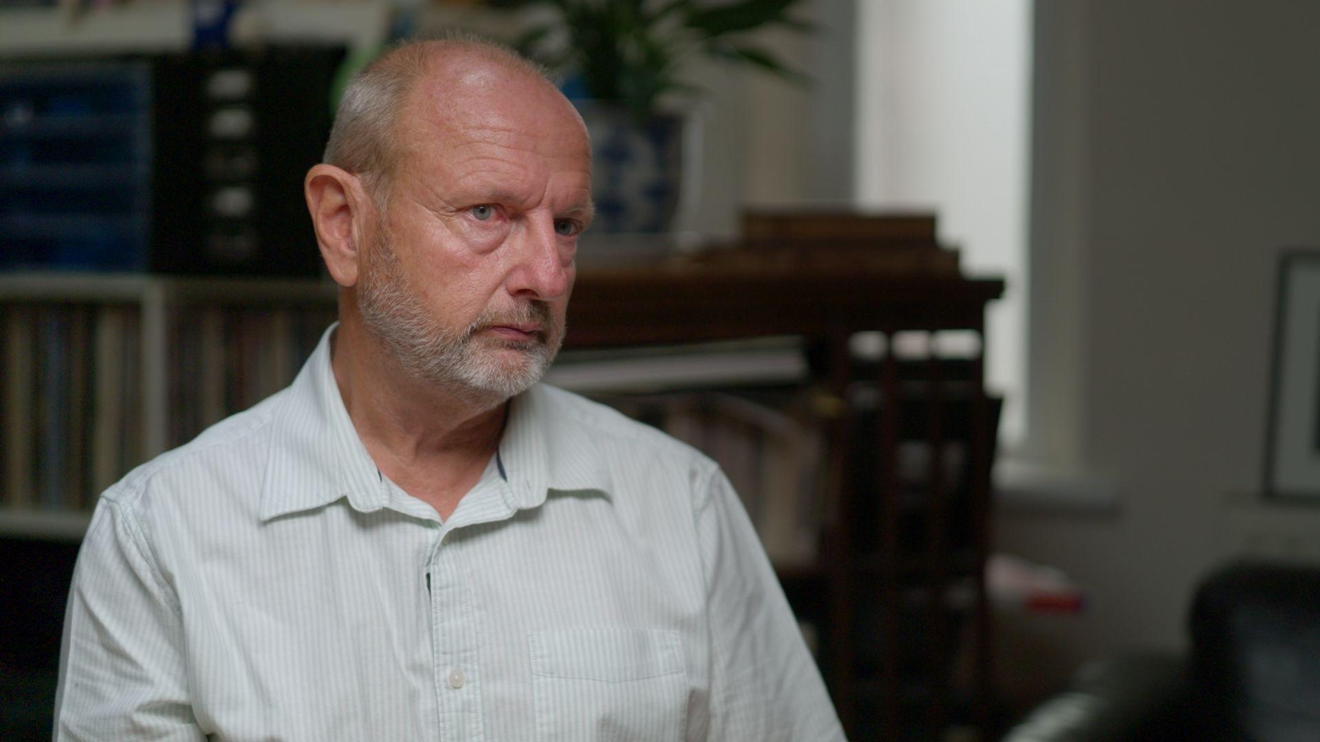 Simon Morton, a man with greying hair at the sides of his head and a trimmed, greying beard, wearing a shirt with pale blue stripes, sits in a room with shelves and plants behind him.