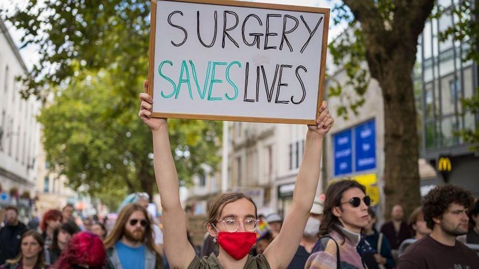 A crowd of people walking down a busy shopping street. The young person at the front of the group has tied-back brown hair and glasses and is wearing a red face mask. Their sign, which they are holding above their head, says: "Surgery saves lives".