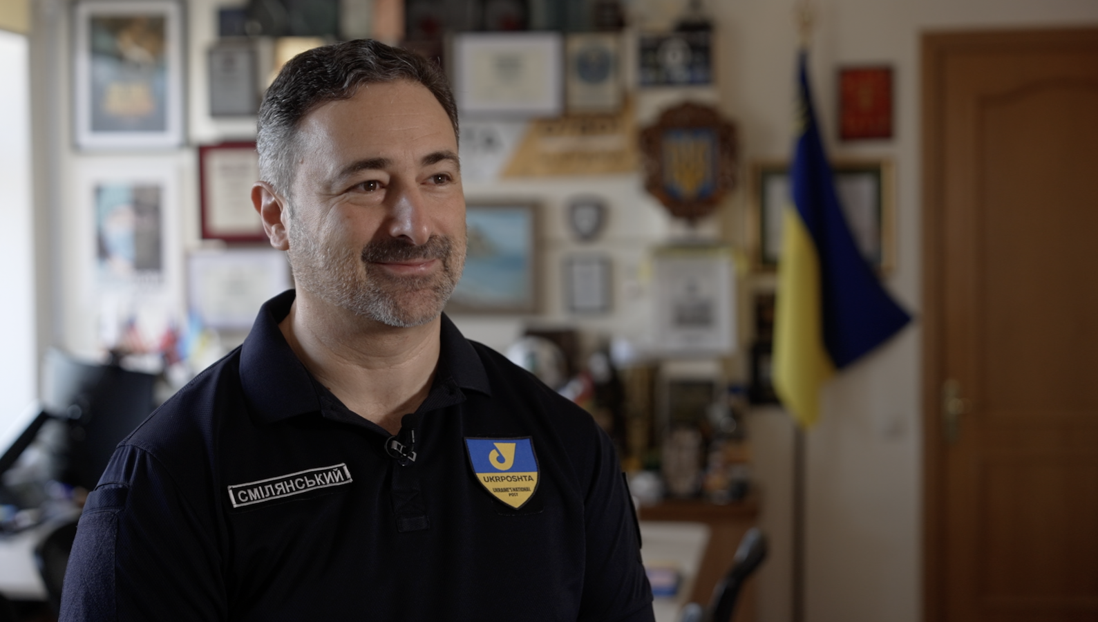 Igor Smelyansky stands in his blue Ukrposhta shirt and smiles at the camera. He has greying dark hair, and wears stubble with a smile