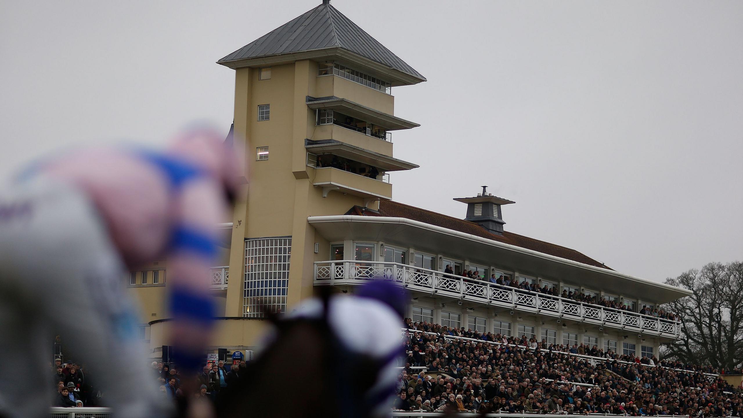 An action shot of a jockey wearing white trousers and a pink and blue striped top, riding a horse at Towcester Racecourse. People are in the stands watching.