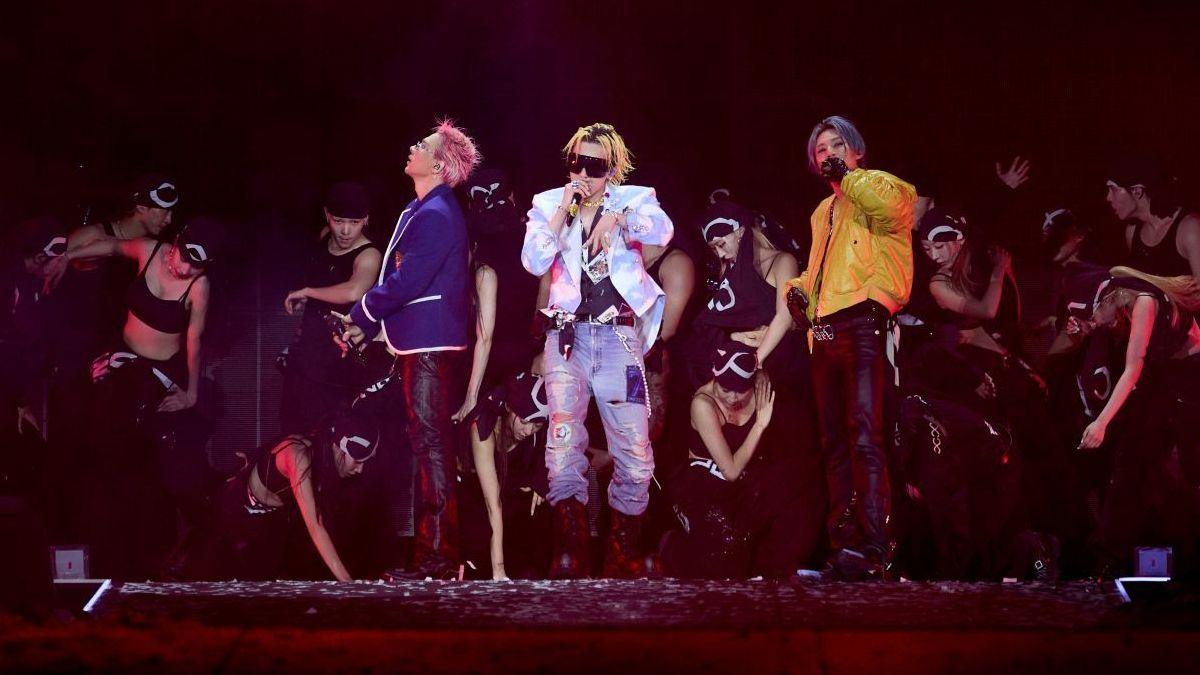 Three performers stand at the centre of a stage holding handheld microphones, surrounded by a group of dancers in black outfits. One performer wears a blue jacket, another wears a white patterned jacket with sunglasses and light-coloured jeans, and the third wears a yellow jacket and dark trousers. Several dancers are positioned crouching or standing around them. The stage is lit with red and purple lighting, with a large dark screen or backdrop behind the group.