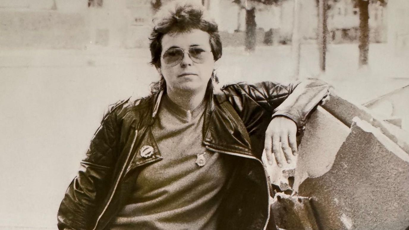A black and white photo of a woman in her late 20s. She is leaning against a skip and has aviator sunglasses on and a black leather biker jacket. She has short hair and a neutral expression on her face.