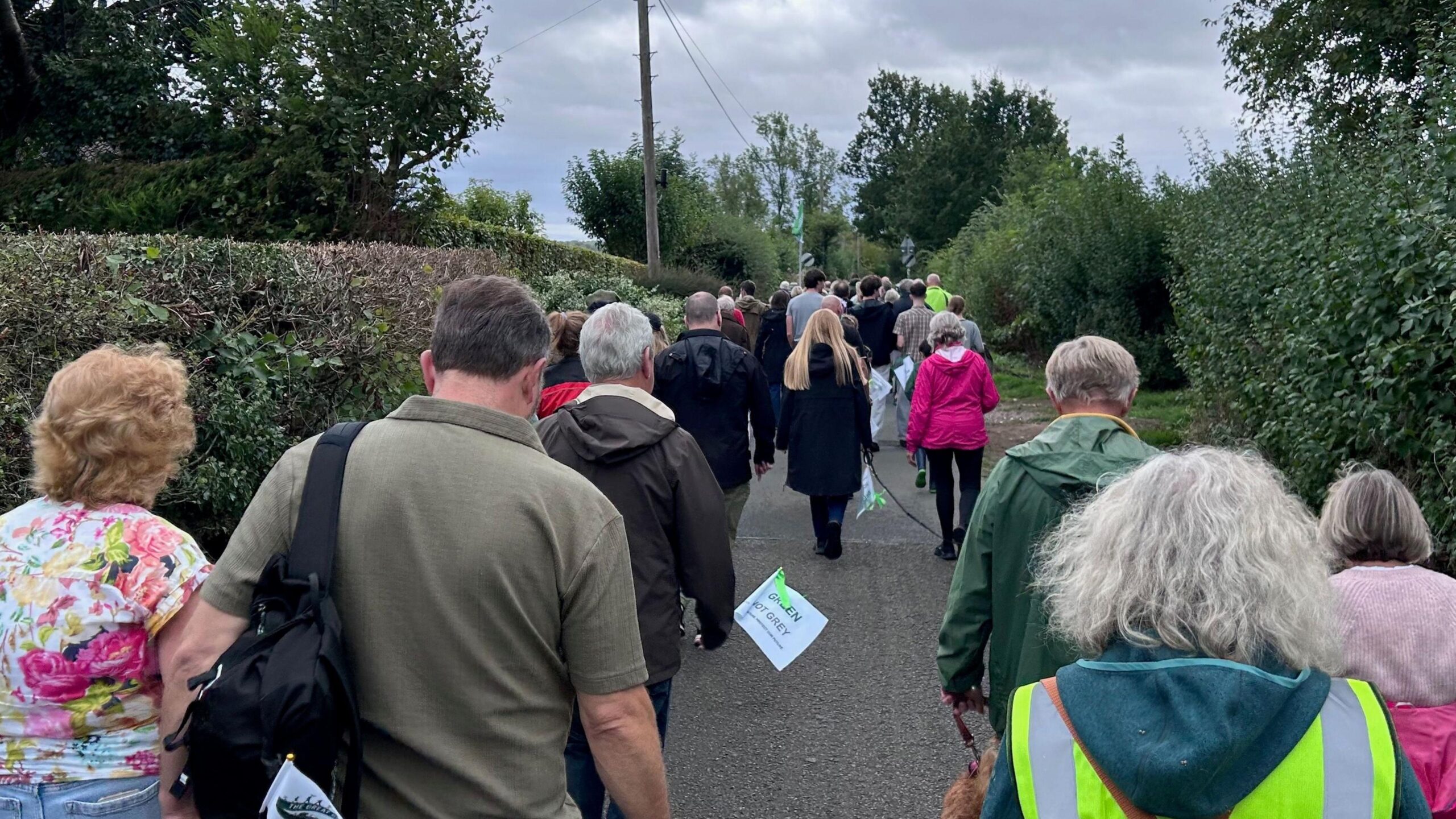 A trail of people walk along a country lane, lots carry white banners saying "Green not Grey"